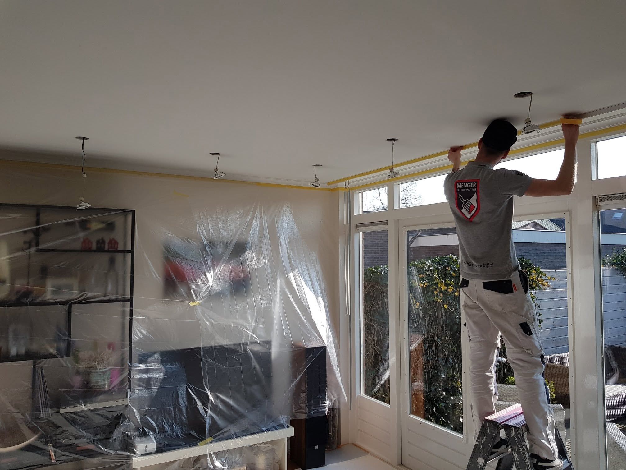 Thijs plakt een plafond af in een woonkamer waar meubels en vloer met folie zijn afgedekt